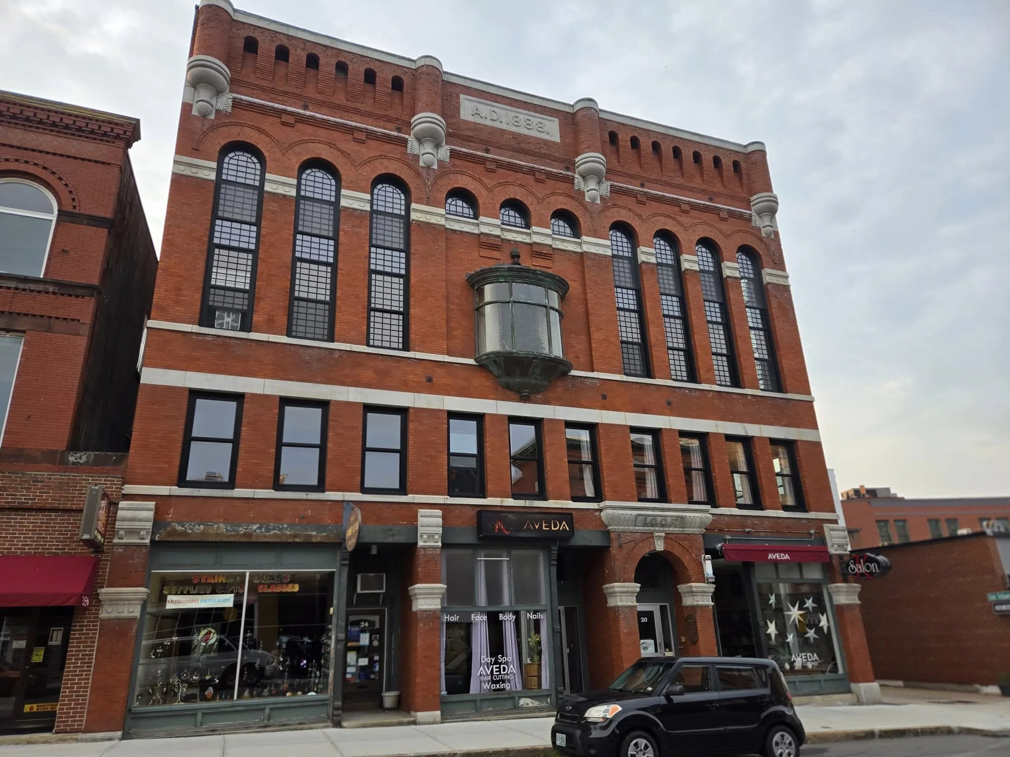 A multi-story red brick building with black window frames and a bay window on the second floor. The storefronts on the ground floor include a hair and nail salon and an Aveda salon. A black car is parked in front of the building.