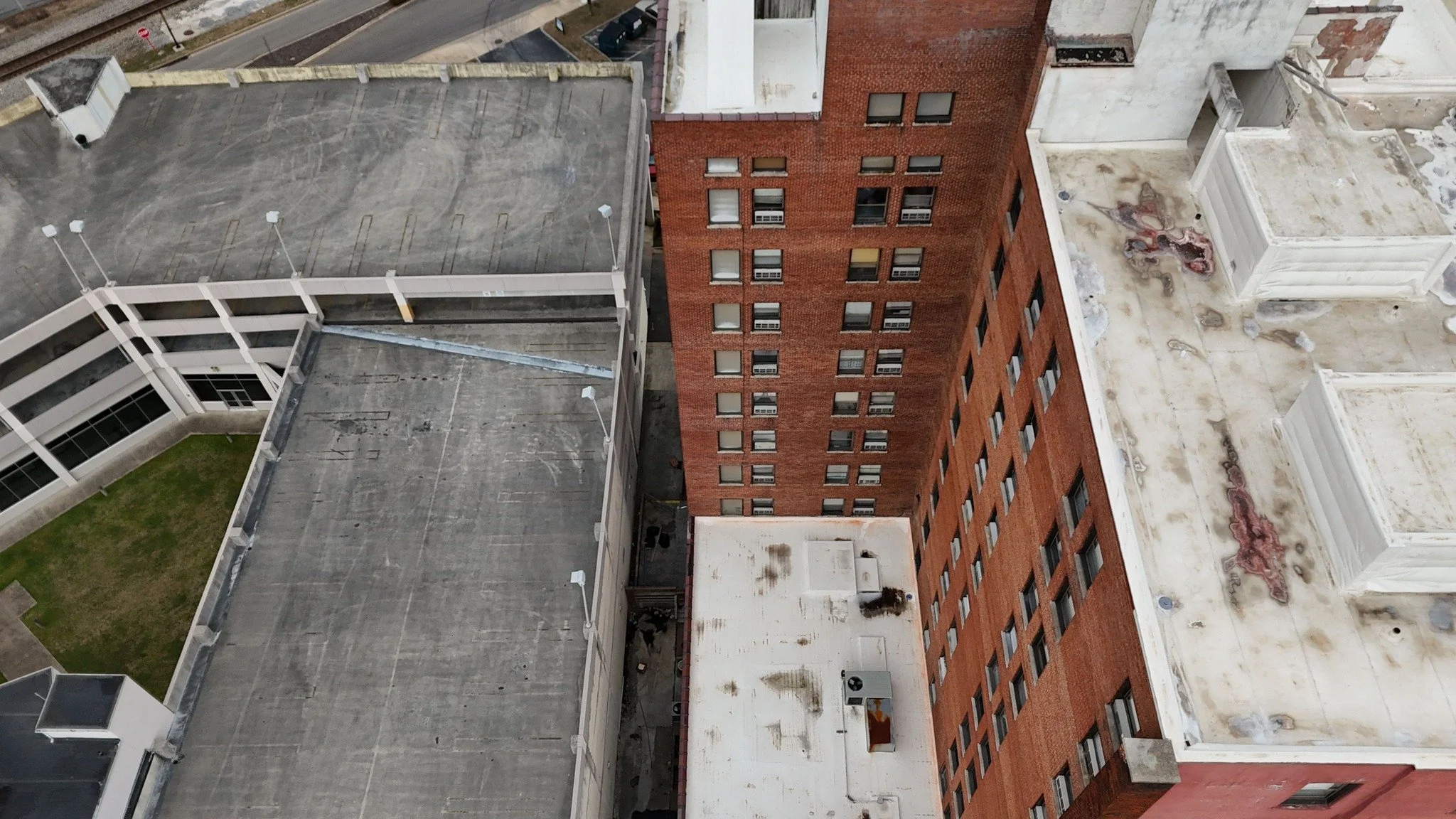 Aerial view of multiple rooftops on a city building, with a red brick building in the center, flat rooftops, some with vents and patches, and a small section of street with parking visible at the top left corner.