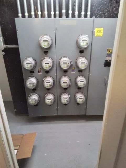 An electrical panel with twelve circular meters or switches arranged in four rows and three columns, located in a utility room with a gray floor and black and beige walls.