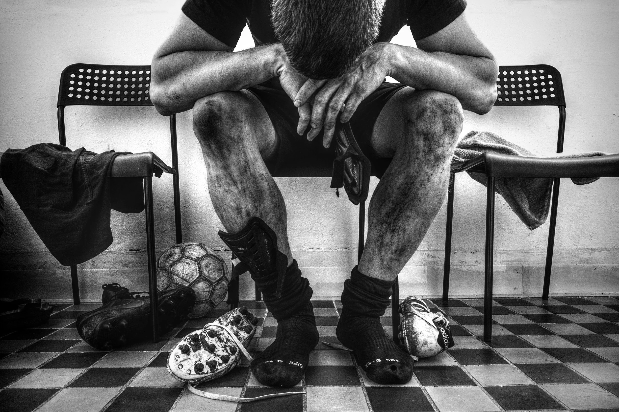 thlete sitting in a locker room after a hard game, leaning forward with head down, surrounded by worn cleats and a soccer ball on a checkered floor, capturing a raw and authentic moment of exhaustion.