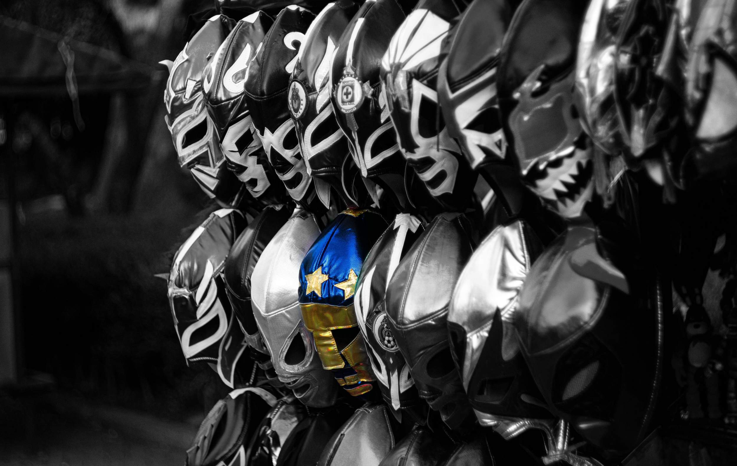 A black and white  photo depicting a display of wrestling masks arranged in rows, featuring a single blue and gold mask standing out from the rest.