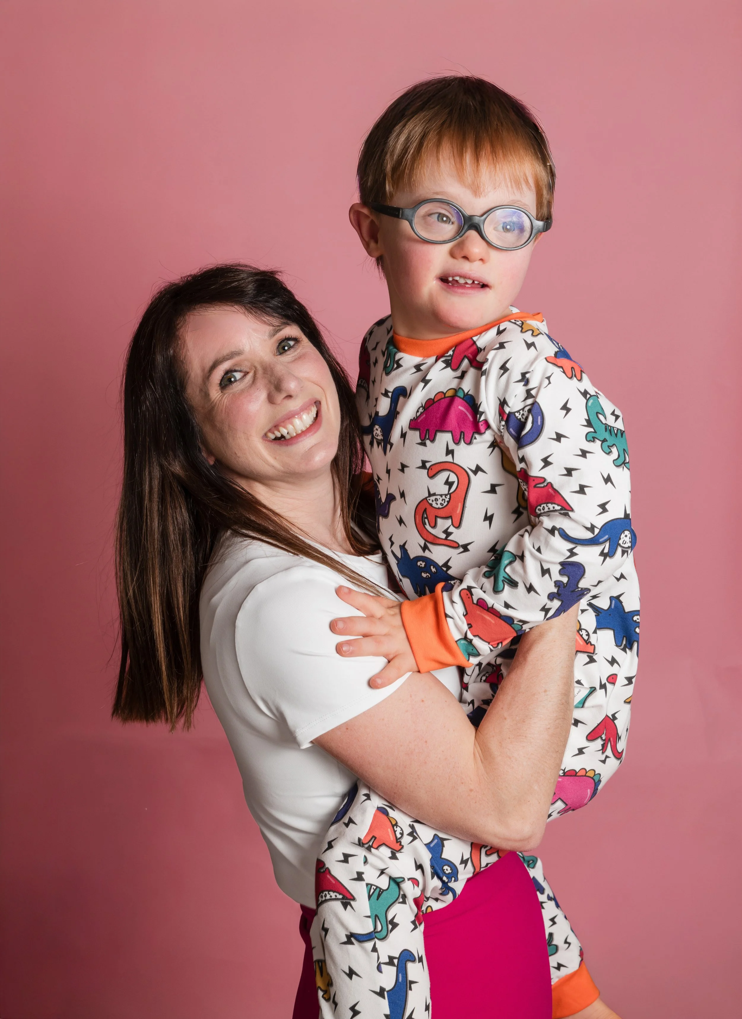 A woman with long dark hair smiling holding a young boy with red hair, glasses, and a colorful dinosaur-print outfit against a pink background.