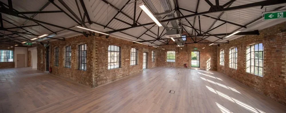 Spacious renovated industrial loft with exposed brick walls, large arched windows, timber flooring, and steel roof trusses