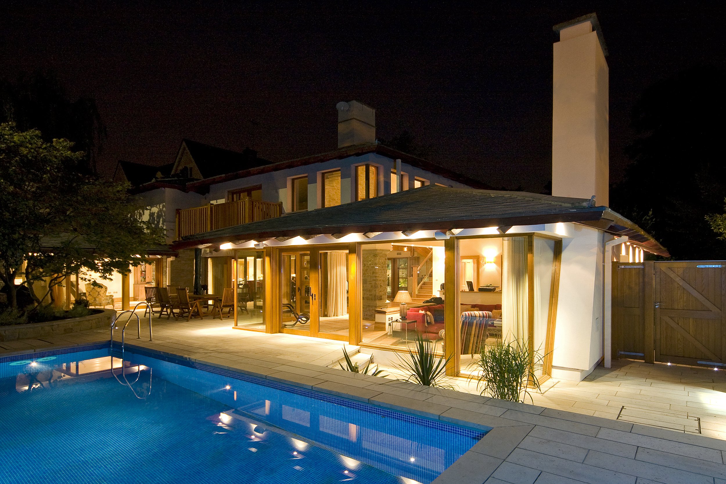 Luxurious modern home illuminated at night with a swimming pool, patio, glass walls, and landscaped garden