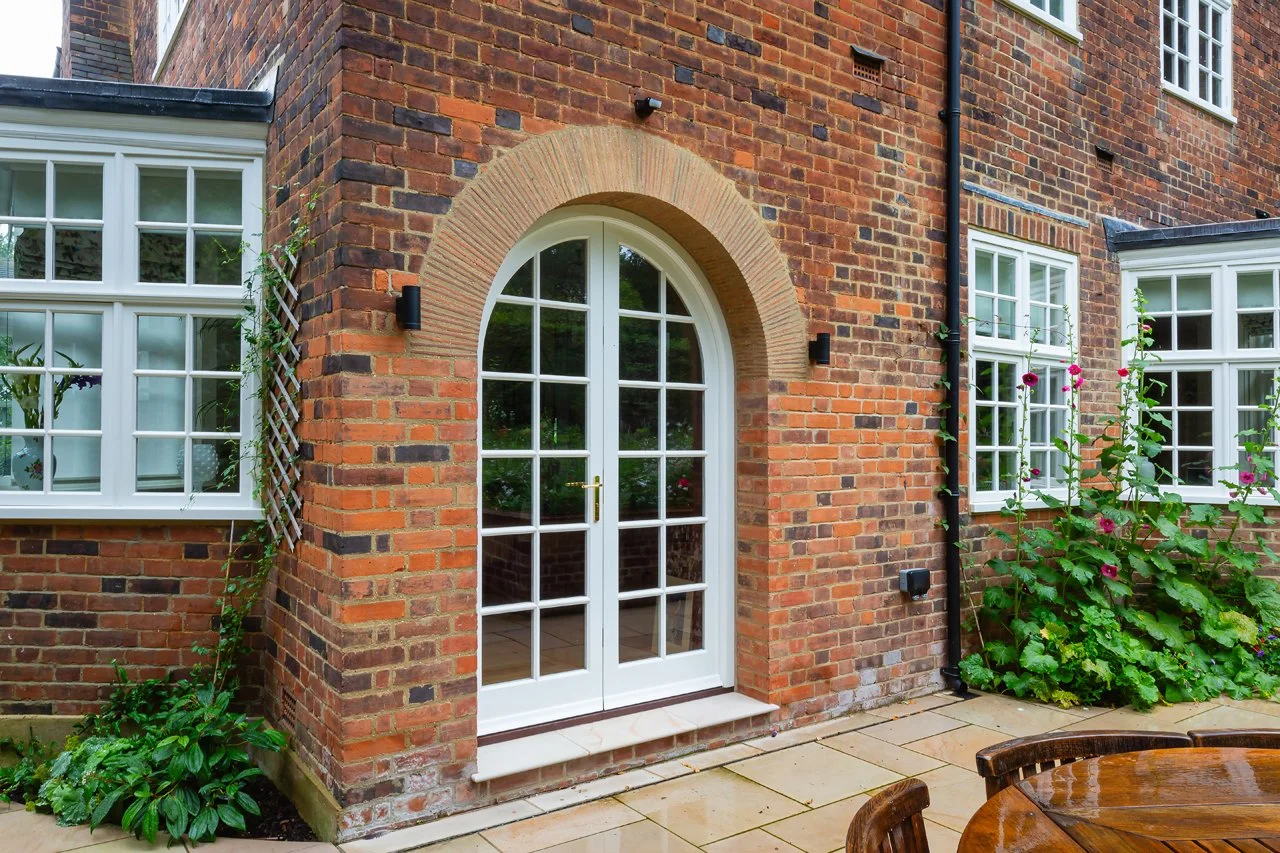 White French doors set in a brick arch on a red brick house, surrounded by windows, climbing plants, and a patio.