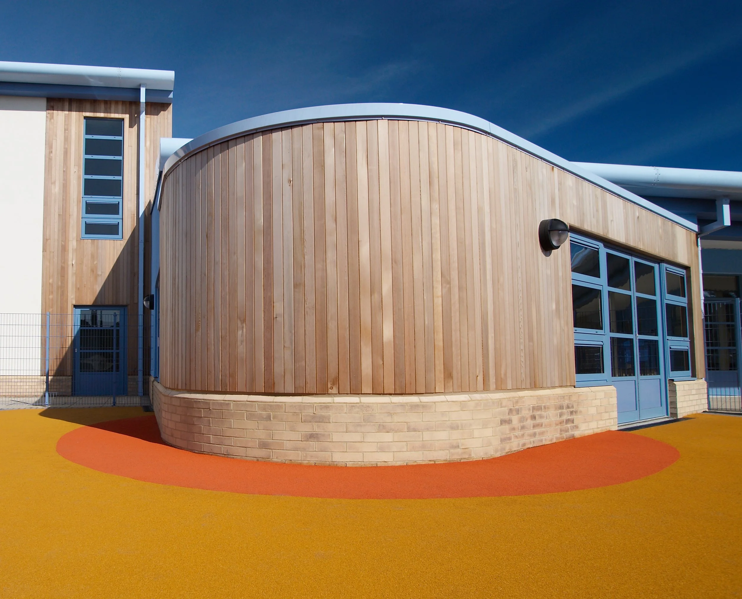 Modern wood-clad building with a curved roof, blue-framed windows and doors, and colorful rubber flooring