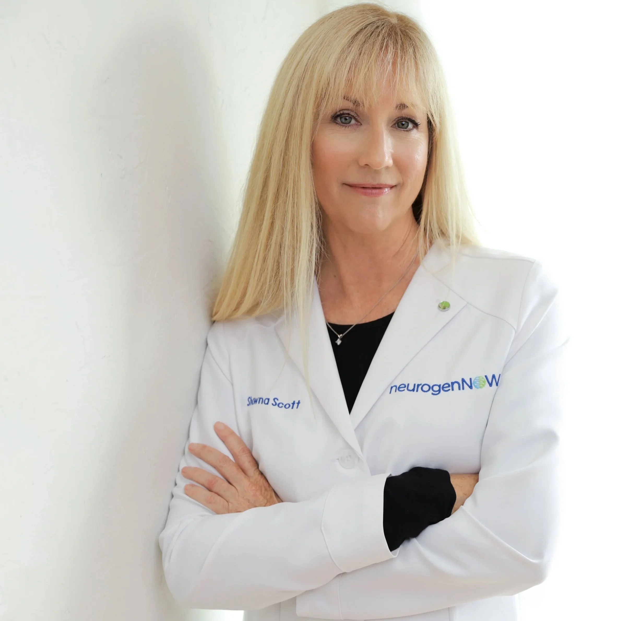 A blonde woman in a white lab coat with 'Shawna Scott' embroidered on it, standing against a white wall with her arms crossed, smiling at the camera.