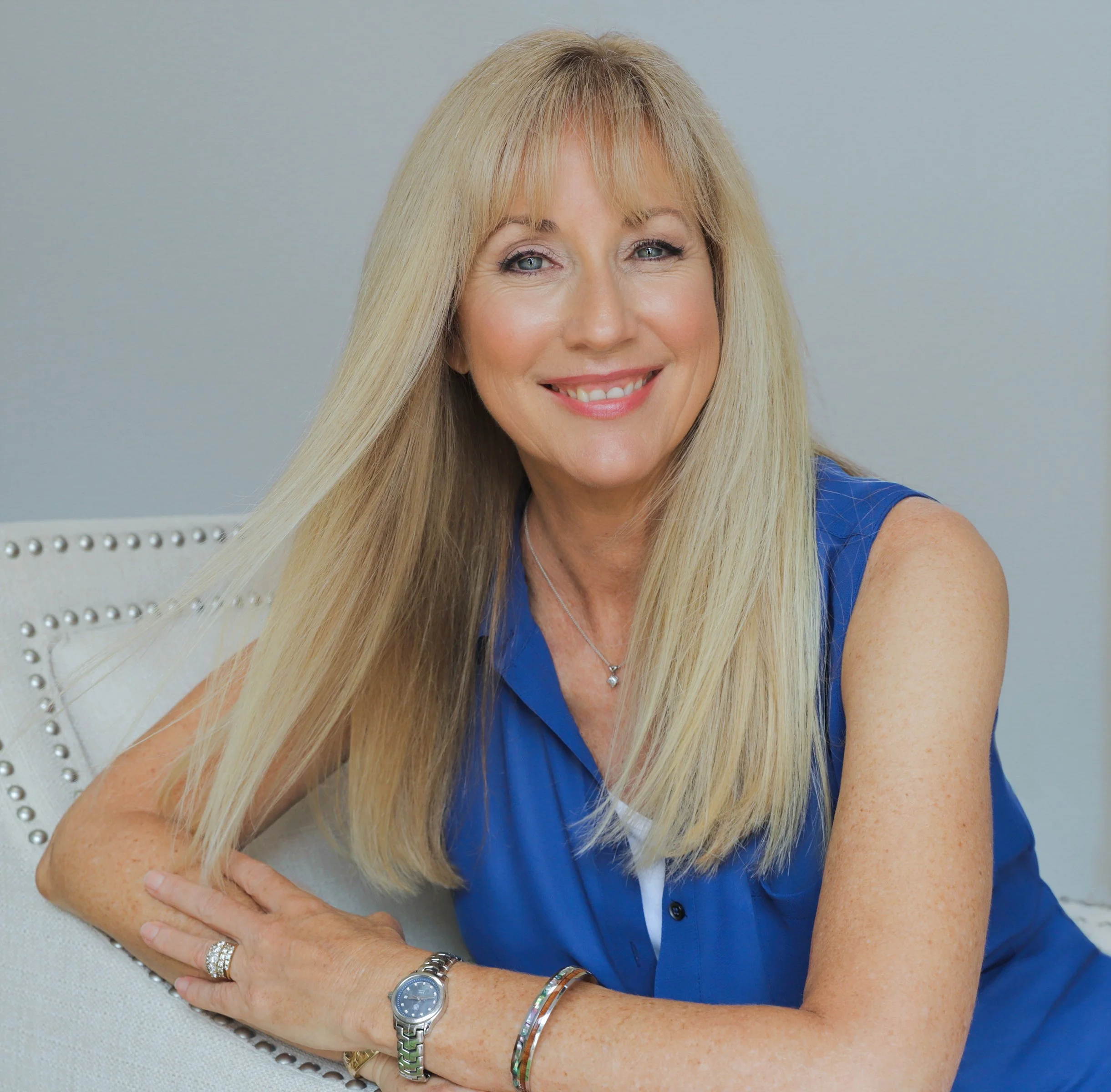 A smiling blonde woman wearing a blue sleeveless shirt, sitting on a light-colored chair with nailhead trim, with long straight hair, jewelry, and a watch, posing against a plain light gray background.