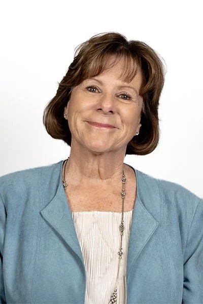 A woman smiling, with dark hair and wearing a blue top, in front of green foliage.