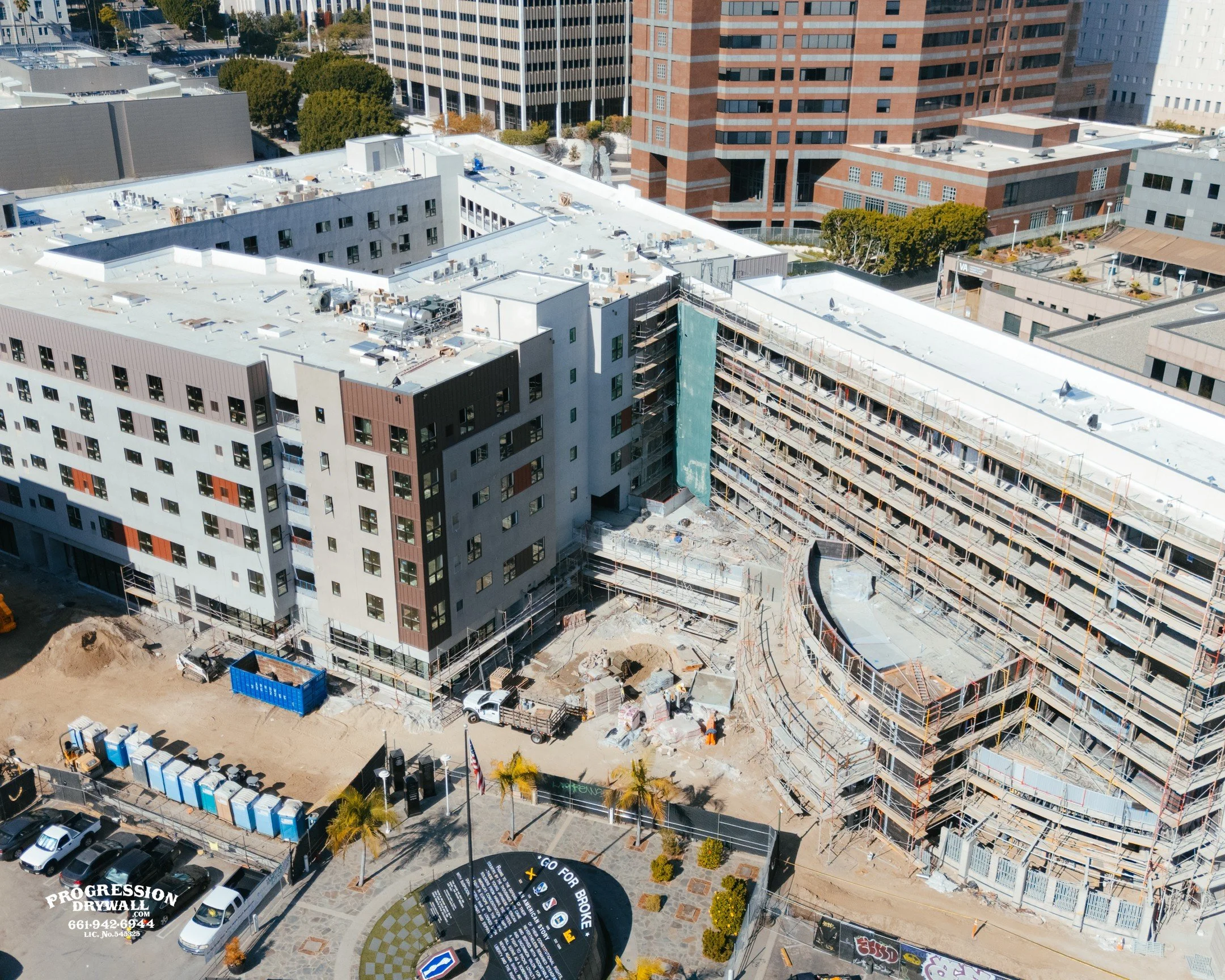 An aerial view of a construction site on a multi-story building in an urban area with surrounding high-rise buildings. Construction scaffolding and equipment are visible, and construction workers are working on the site.