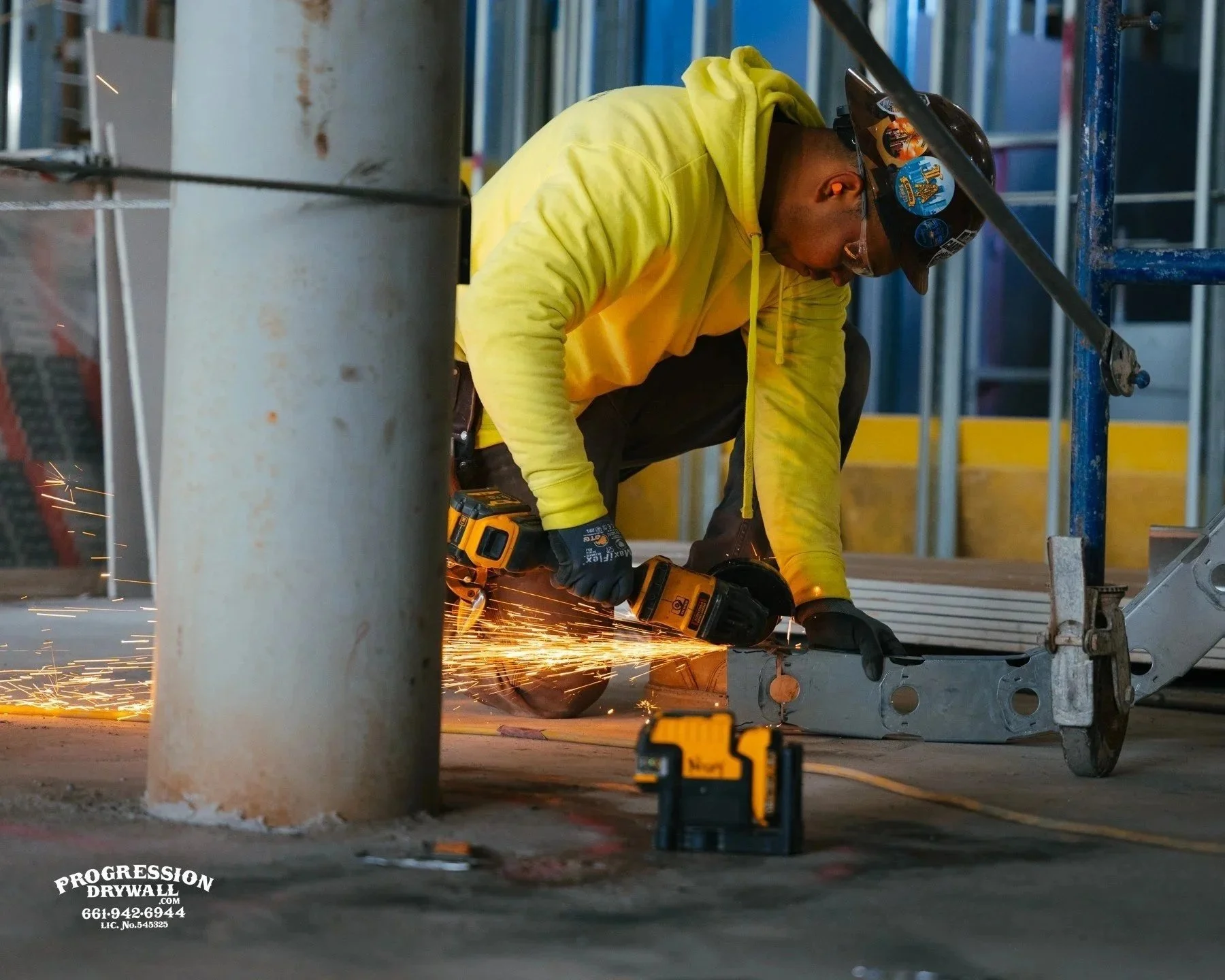A construction worker wearing a yellow hoodie, safety glasses, and a hard hat, is using a power drill to fasten a metal piece, with sparks flying from the drill. The worker is crouched on the ground amid construction scaffolding.