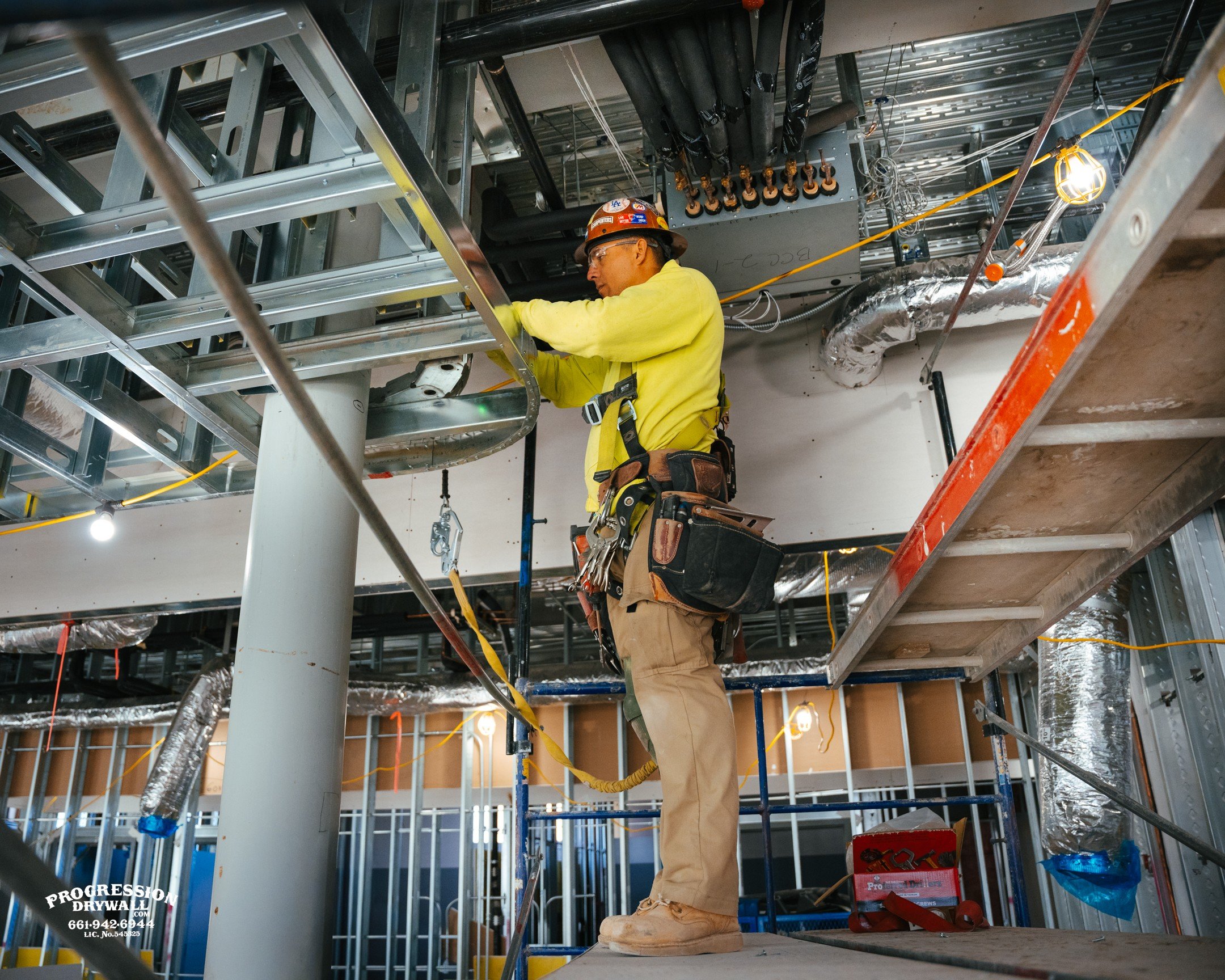 Construction worker in yellow shirt and safety harness working on metal framing and ductwork inside a building under construction.