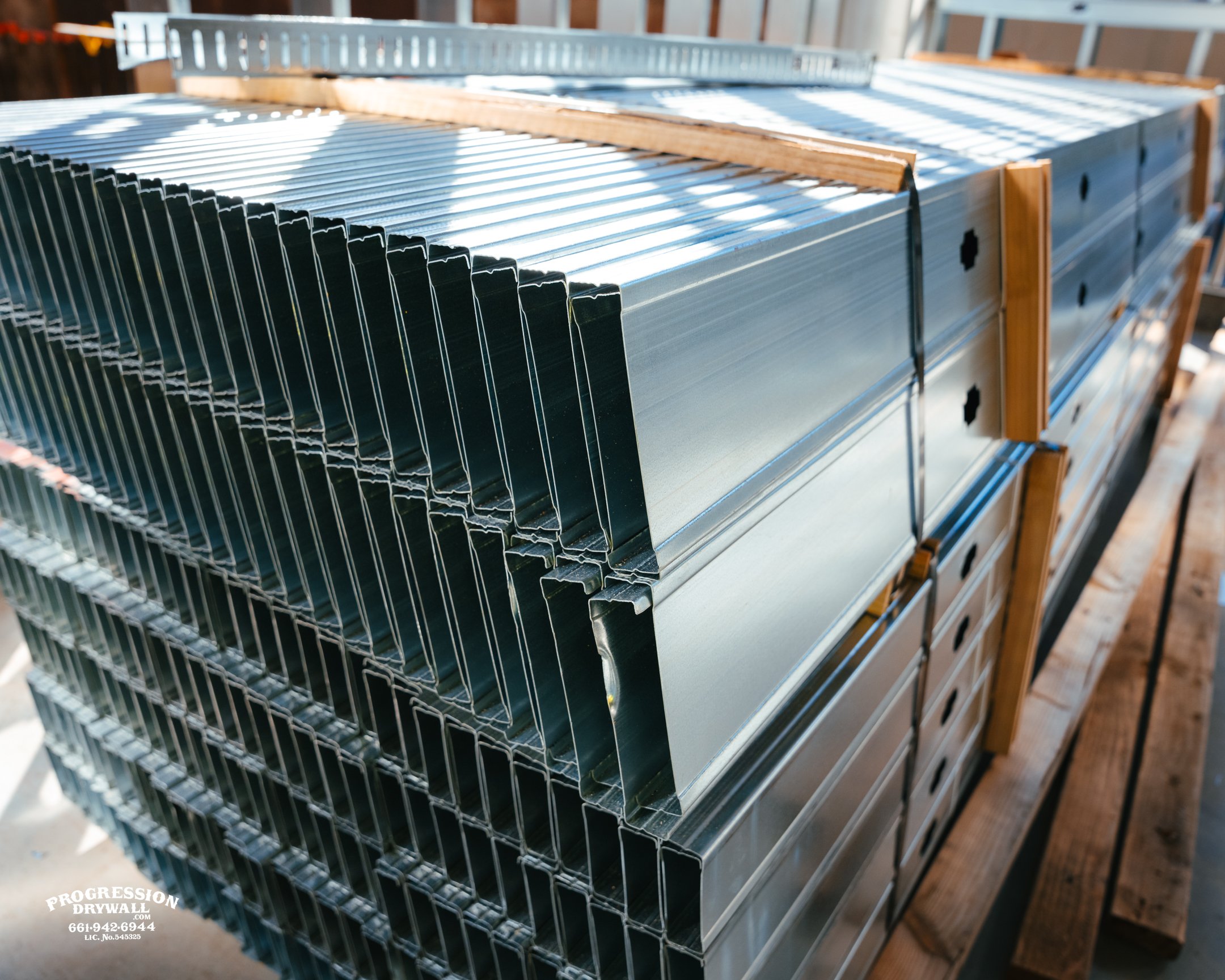 A stack of metal drywall corner beads on a construction site, with ductwork and wooden framing in the background.
