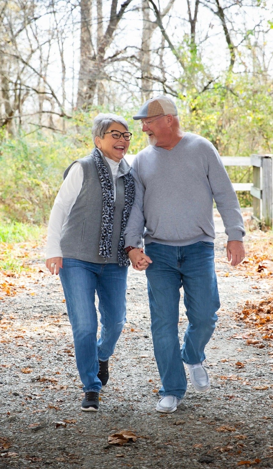An elderly couple walking hand in hand on a wooded path, smiling and enjoying each other's company.
