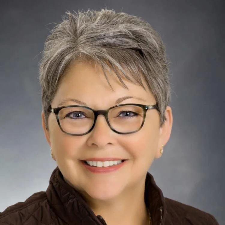 Portrait of a smiling mature woman with short gray hair, wearing glasses and a brown jacket against a dark gradient background.