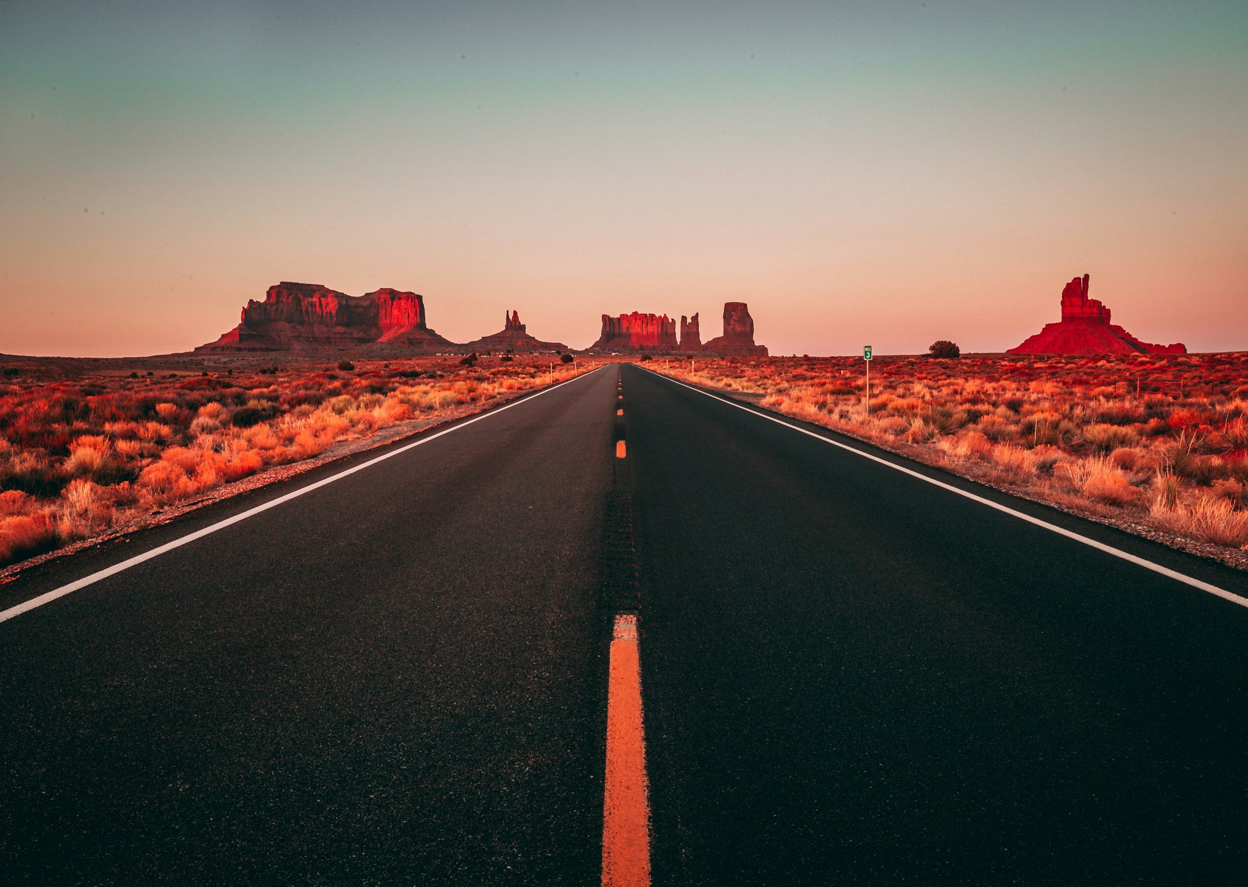 A straight desert highway leading towards the rock formations in Monument Valley during sunset or sunrise, with a clear sky and reddish glow on the rocks.