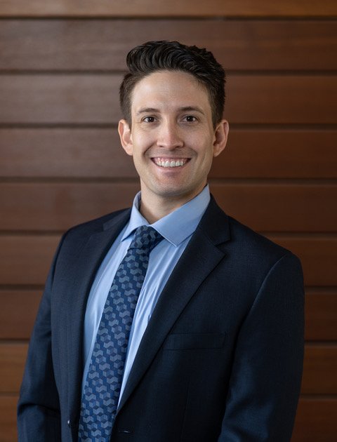 A young man in a dark suit, light blue shirt, and patterned blue tie, smiling, standing in front of a wooden wall.