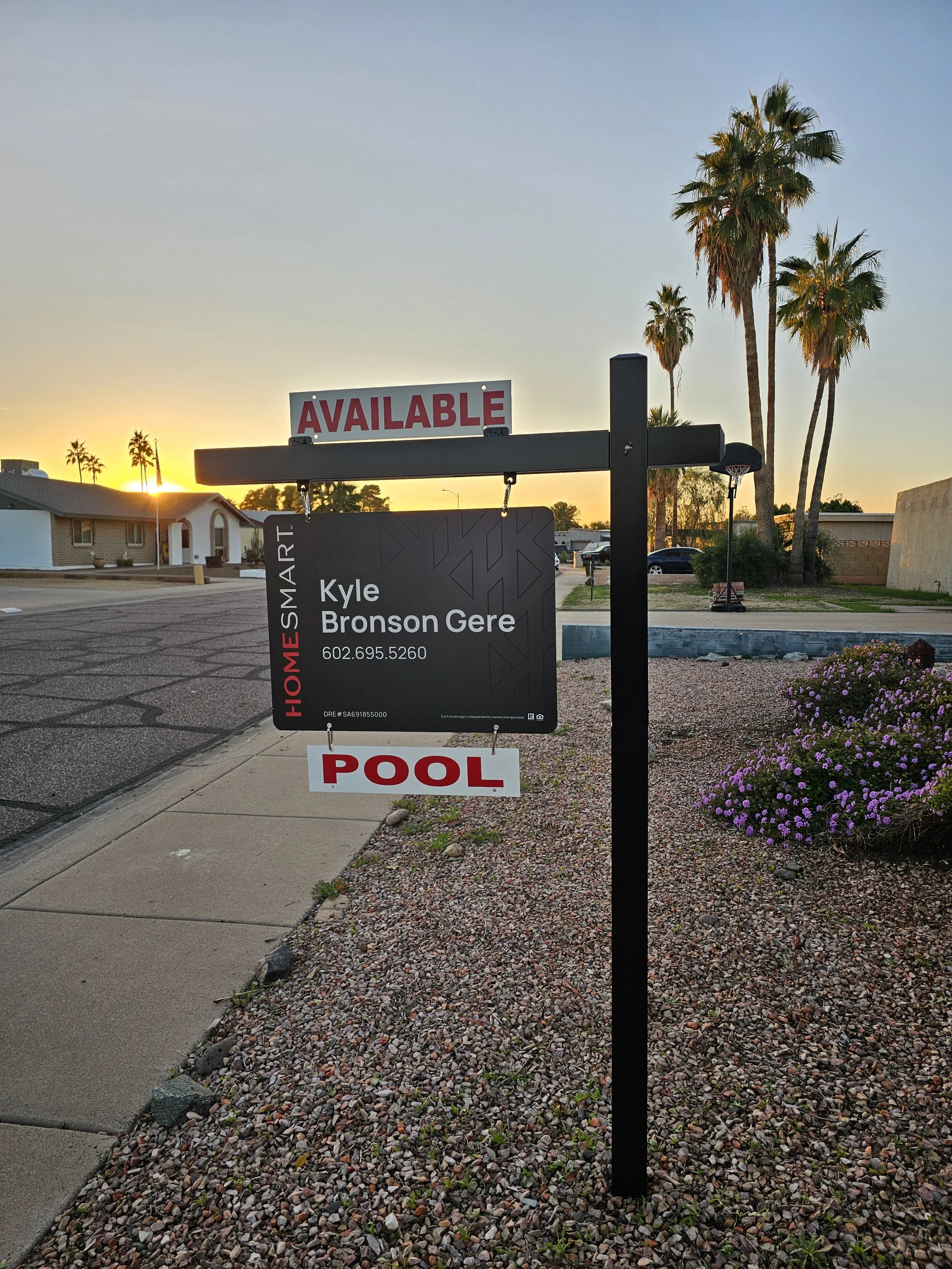 Real estate sign in front of a house with a sunset in the background, palm trees, and a gravel yard with purple flowers.