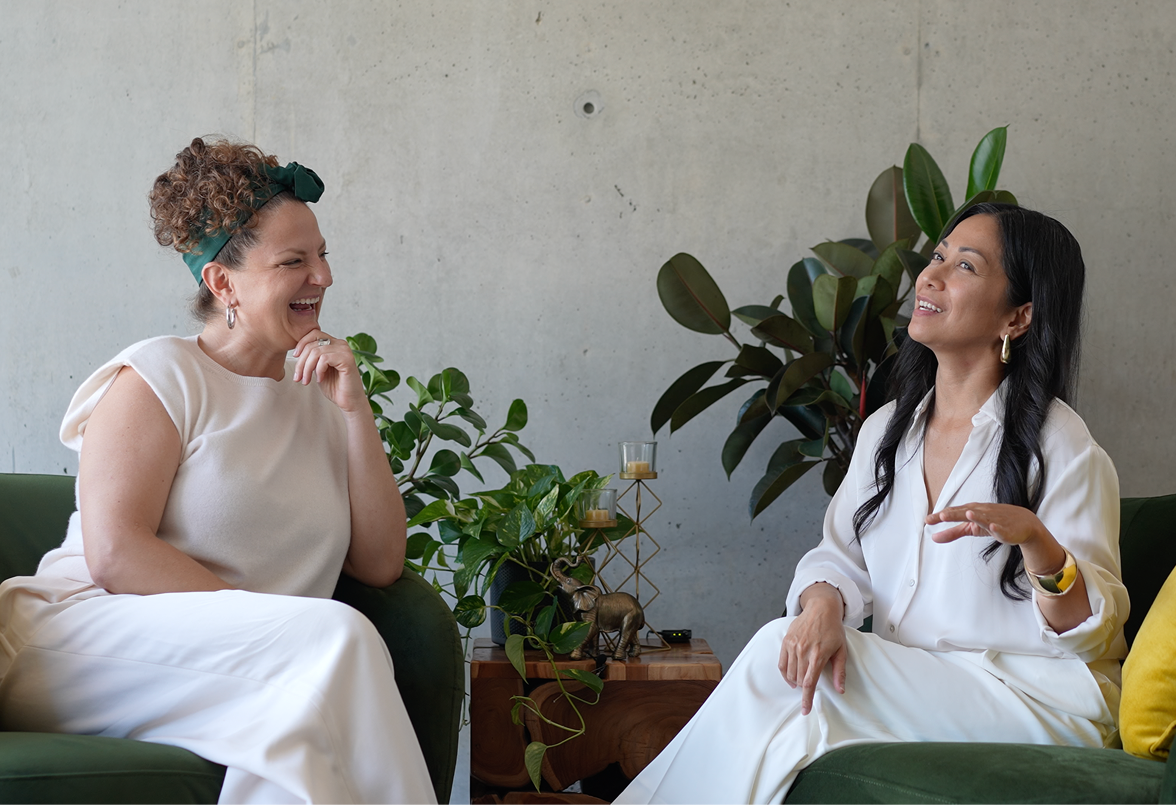 Two women sitting on green sofas, laughing and talking in a modern, minimalist living room with plants and decorative items on a wooden side table.