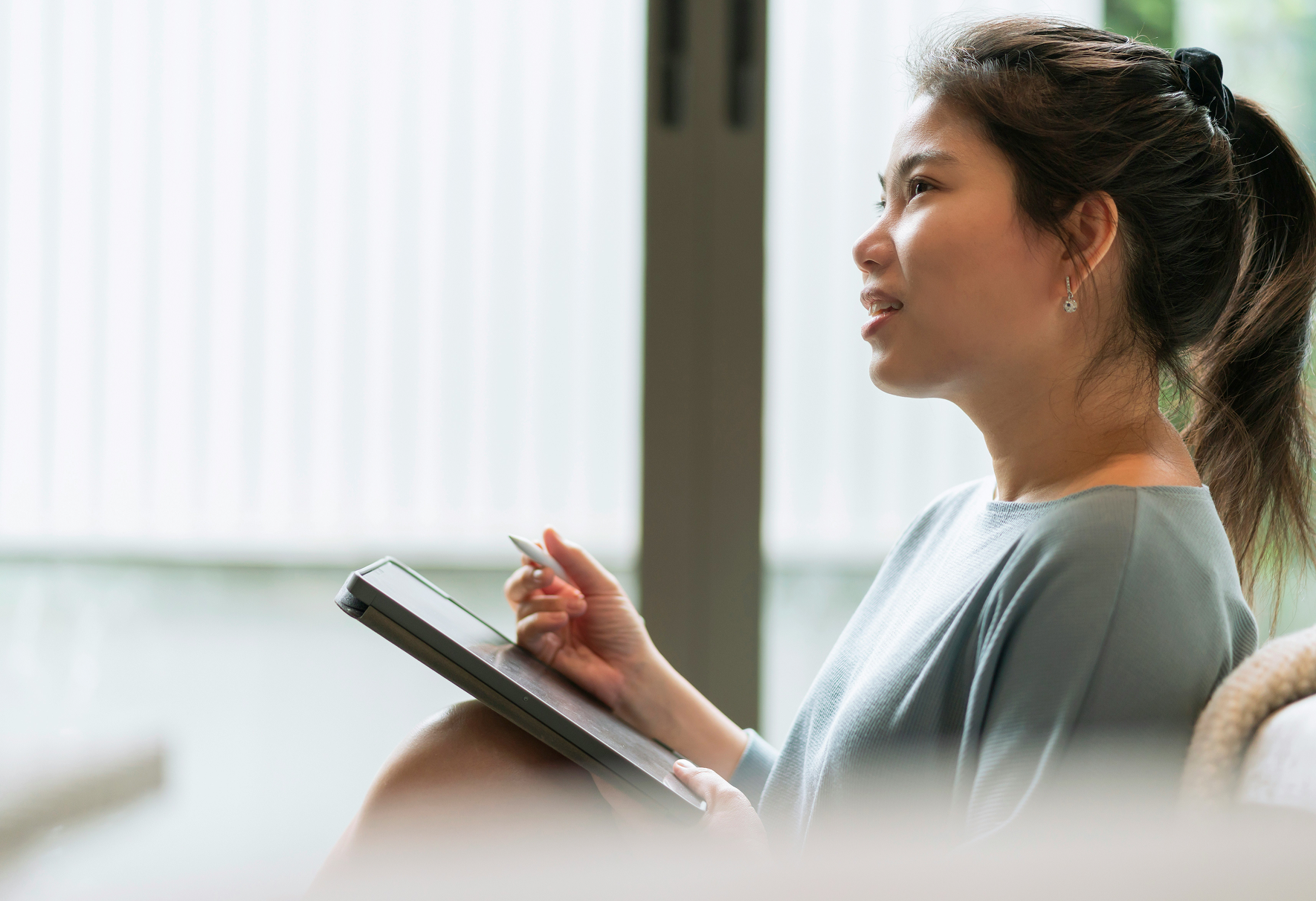 A woman with dark hair tied back in a ponytail, wearing earrings and a gray top, is sitting next to a window, lookng to the side while holding a digital tablet and stylus.