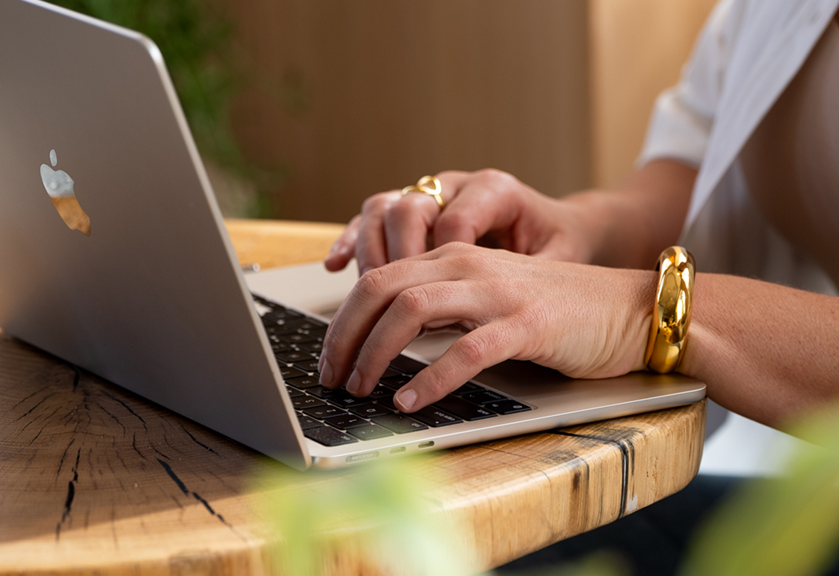 Person typing on a silver MacBook with a wooden table and green plants in the background.