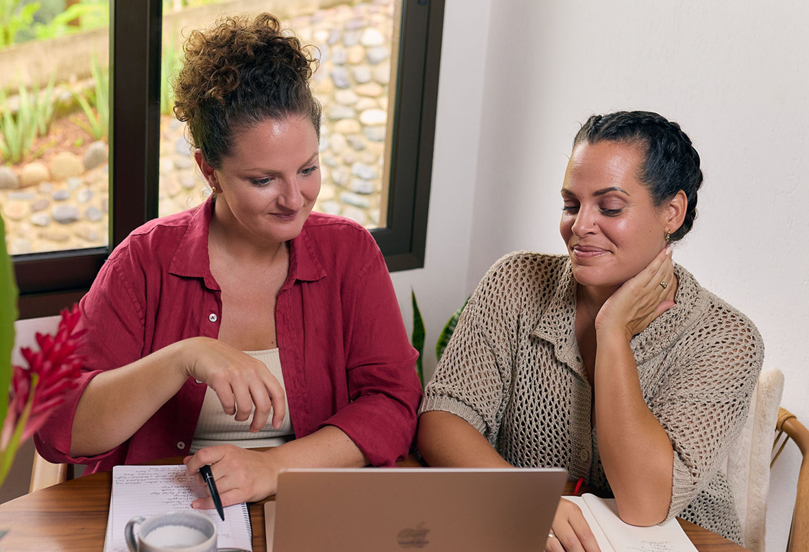 Two women sitting at a table looking at a laptop, one pointing at the screen.