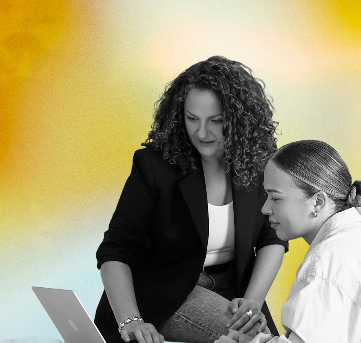 Two women, one with curly hair and one with straight hair tied back, are engaged in a discussion over a laptop in a professional setting with a colorful blurred background.