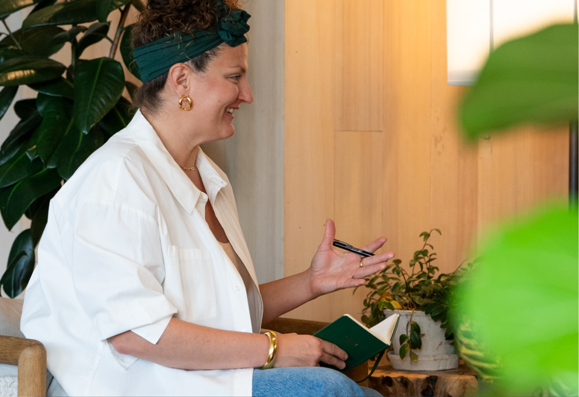 woman sitting on a chair, smiling, wearing a white shirt and blue headband, surrounded by green plants, holding a pen and a notebook.