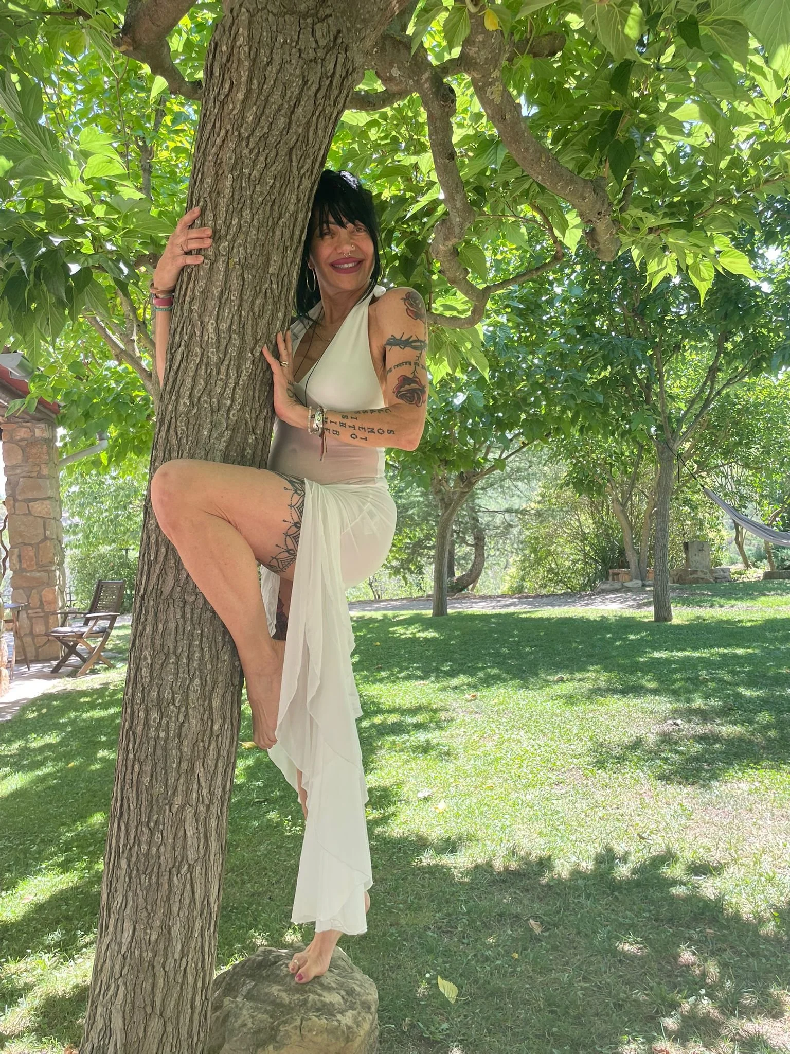 A woman with tattoos, wearing a white dress, standing on a rock with one foot on a tree root, hugging a tree in a park with green trees and grass, and a stone structure in the background.