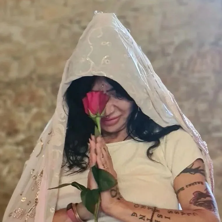 A woman with dark hair, tattoos on her arms, and glasses holding a pink rose with a veil on her head, smiling gently with her eyes closed.
