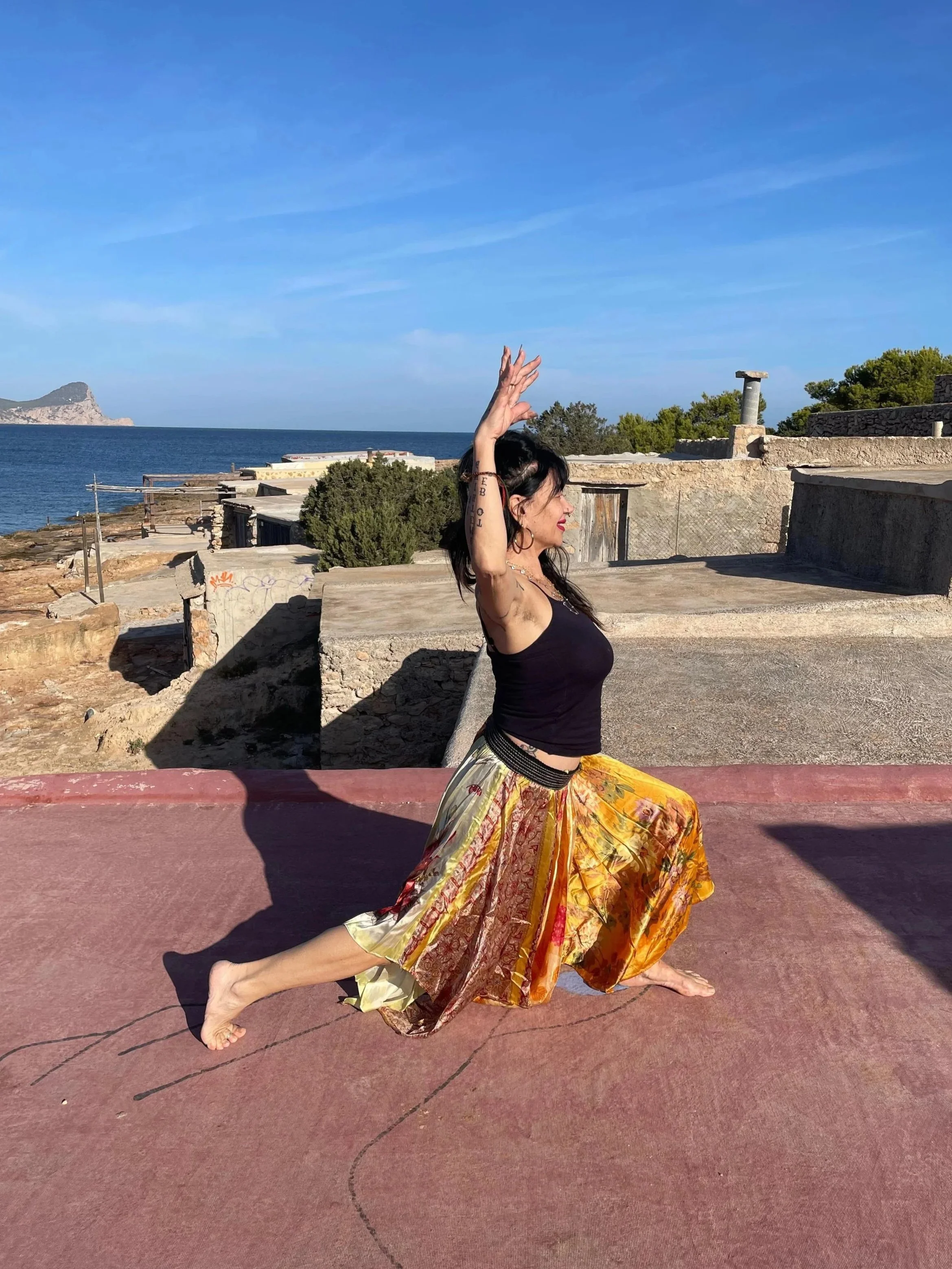 A woman practicing yoga outdoors on a pink surface near the coast, with the ocean and mountains in the background, wearing a black tank top and colorful skirt, holding a yoga pose with arms raised.