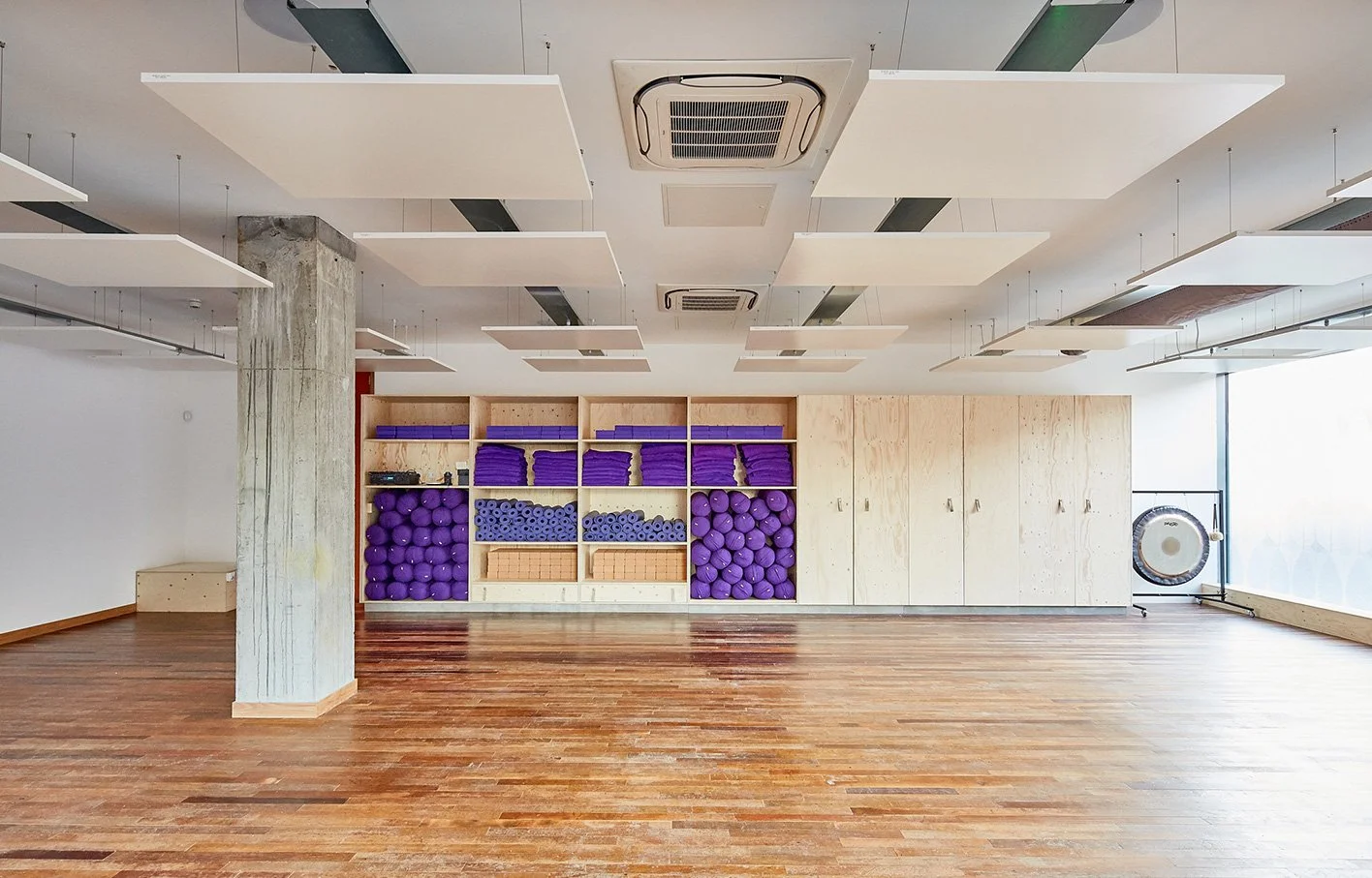 Empty fitness studio with wooden flooring, storage shelves with purple exercise equipment, and large window at the right.