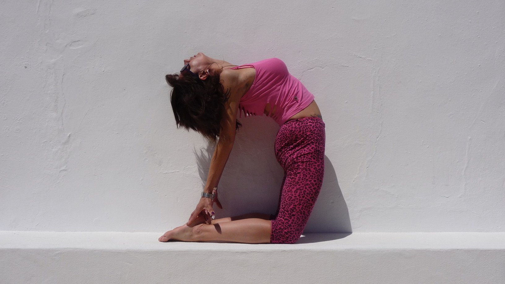 Woman in pink tank top and pink leopard print leggings practicing yoga in a kneeling backbend pose against a plain white wall.