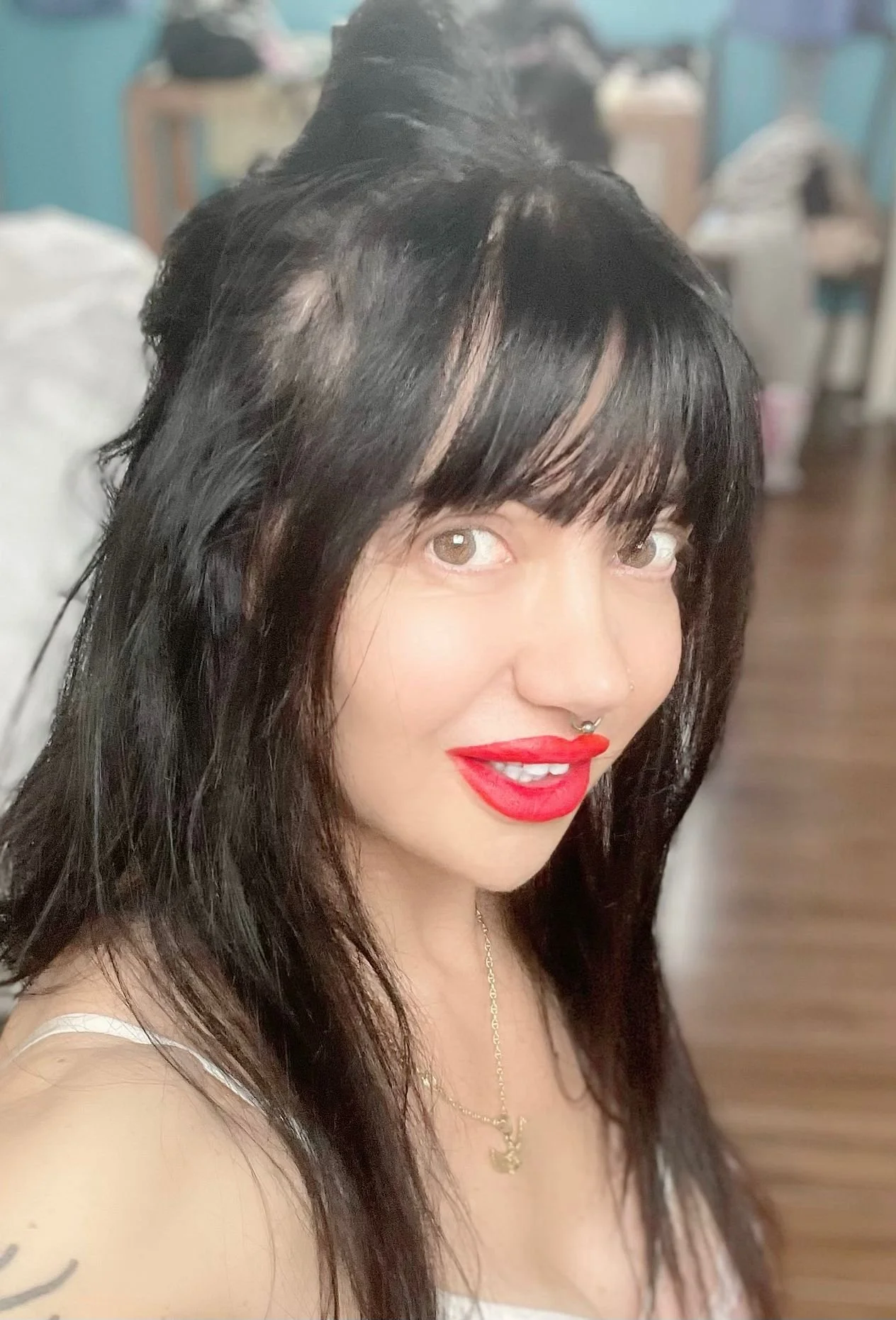 A woman with dark wavy hair, red lipstick, a septum piercing, and a gold necklace, taking a selfie in a room with wooden flooring and furniture in the background.
