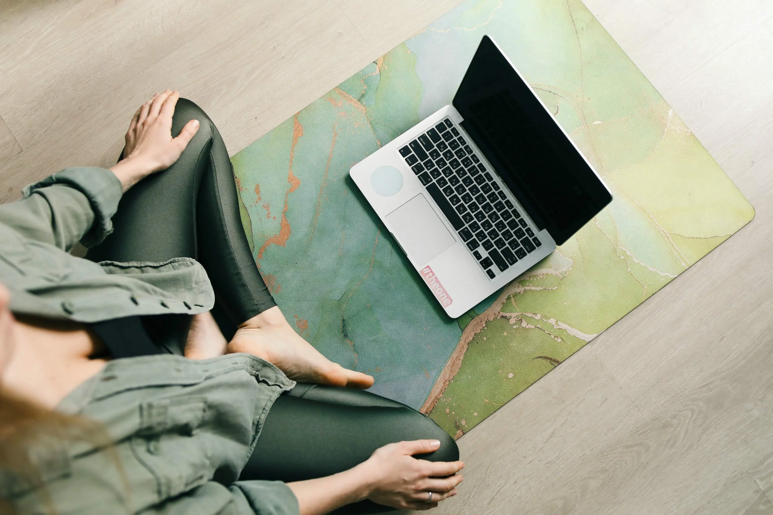 Person sitting on the floor with legs crossed, wearing a green jacket and black pants, next to a colorful rug, with a silver laptop on the rug.