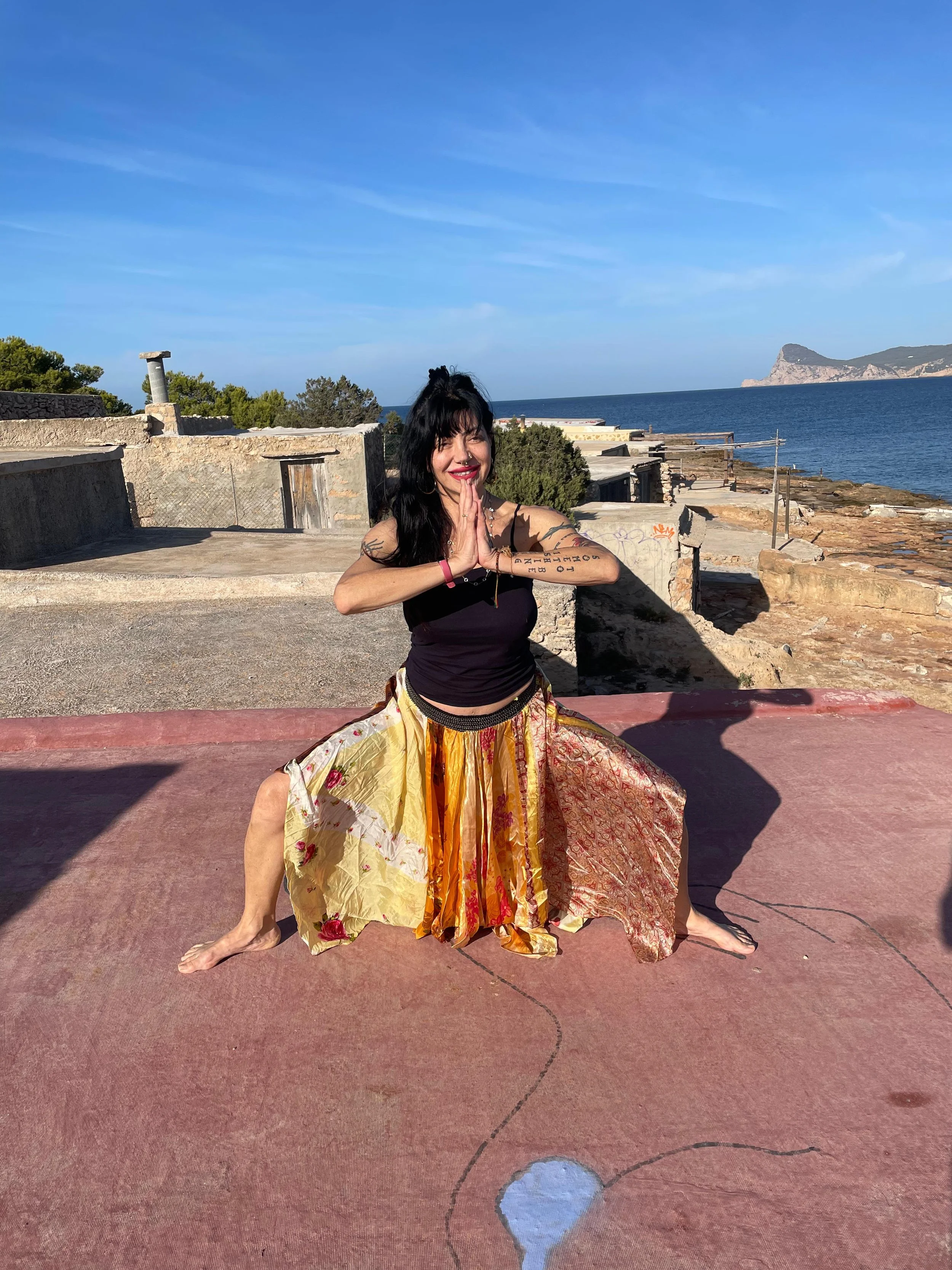 Woman practicing yoga outdoors on a pinkish concrete surface near the ocean, with old buildings and rocky coastline in the background.
