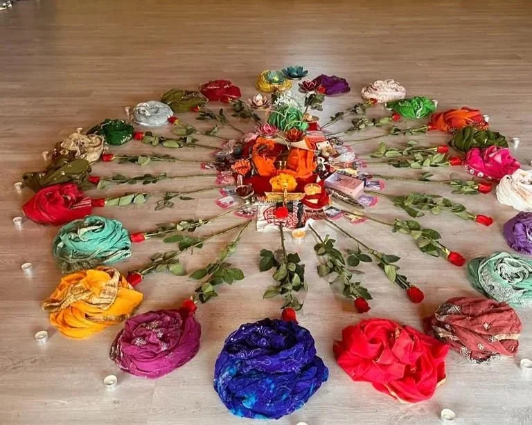 Colorful fabric flowers arranged in a circular pattern on a wooden floor, with small decorative items and leaves connecting them.