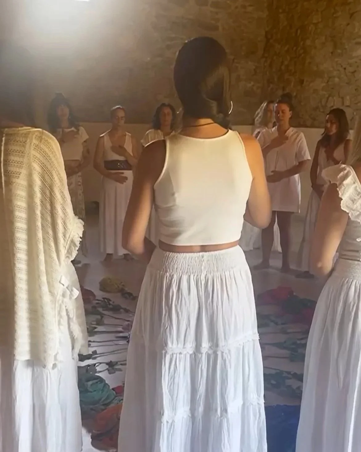 Group of women in white dresses standing in a circle in a rustic room with brick walls, with clothes and shoes scattered on the floor.