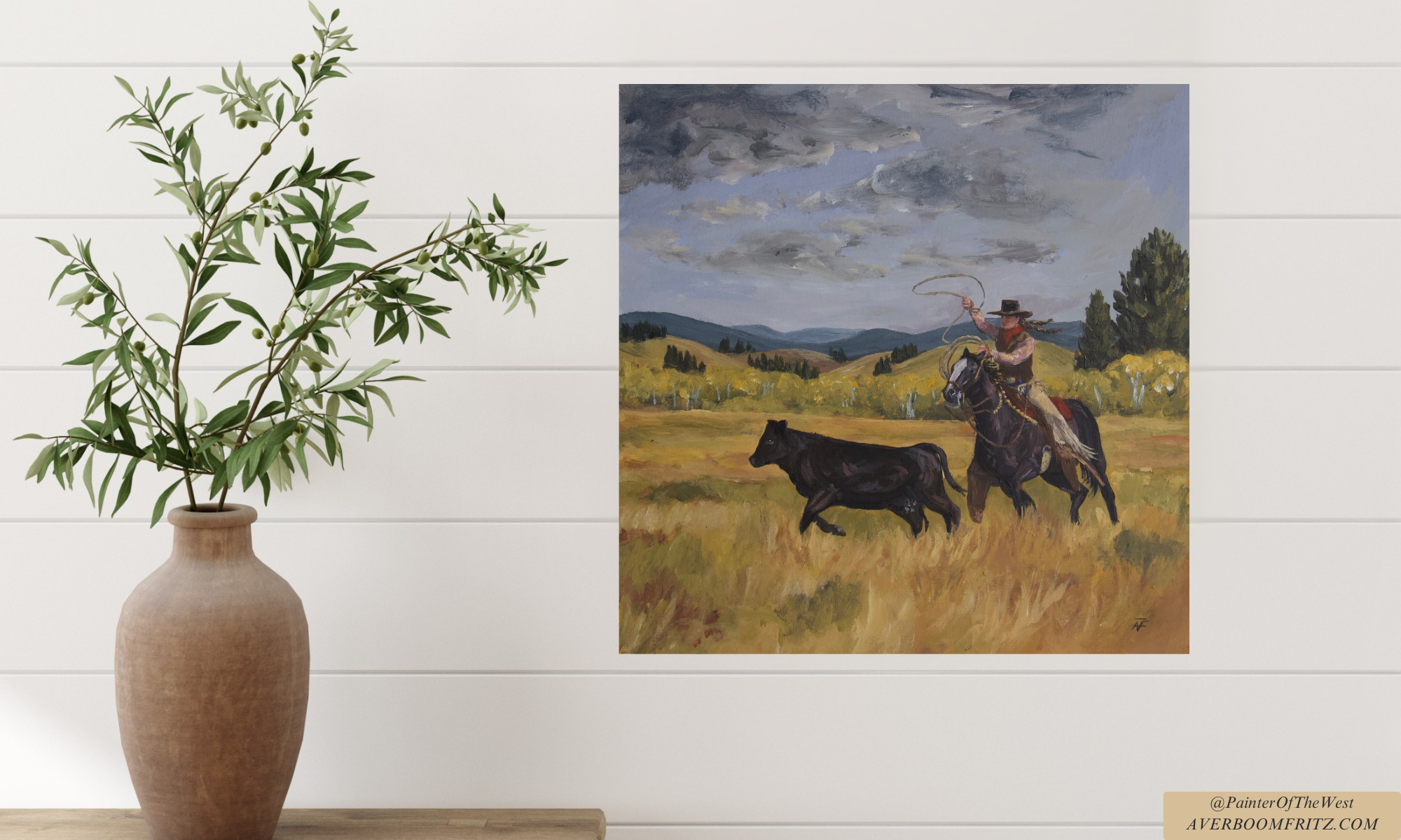 A framed painting of a cowgirl on horseback about to rope a cow in a rural landscape with blue sky and clouds, hung on a white wall next to a large potted plant with green leaves.