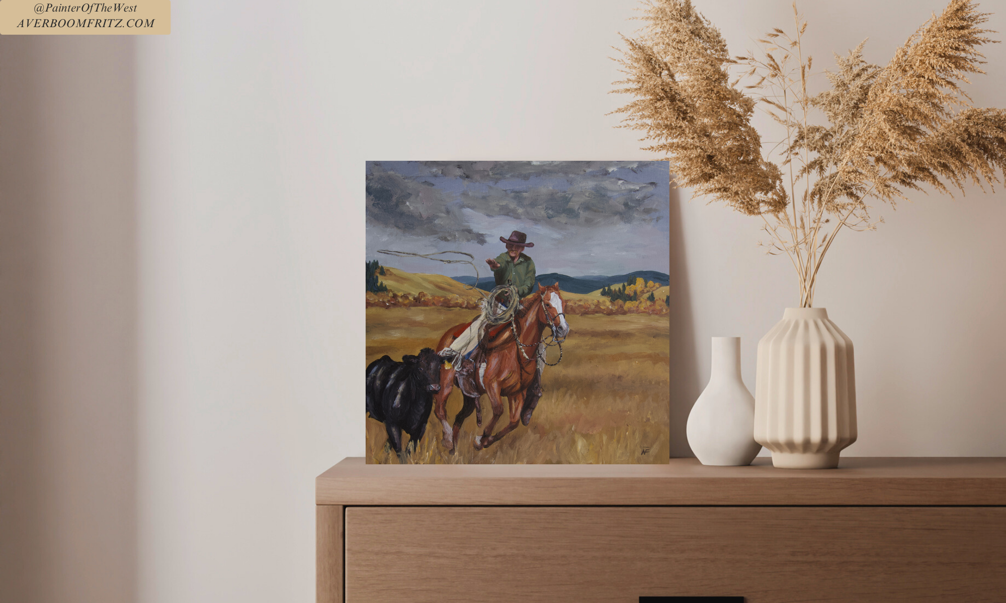 A painting of a cowboy on horseback roping a cow in a grassy field with rolling hills and cloudy sky in the background, placed on a wooden surface next to tall dried pampas grass in a ribbed cream-colored vase and a small white vase.