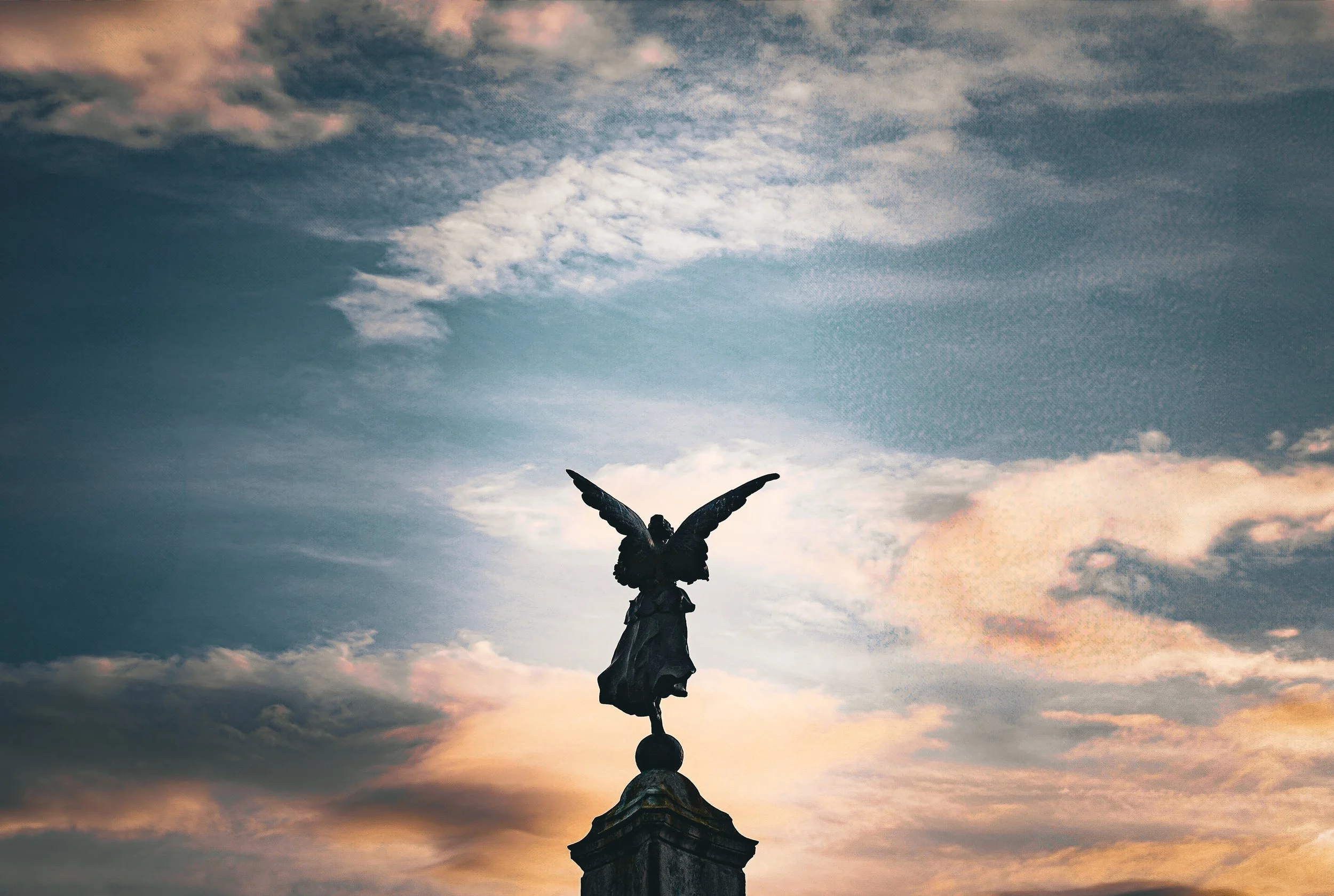A sculpture of an angel standing on a pedestal with wings spread, set against a sky with clouds at sunset.