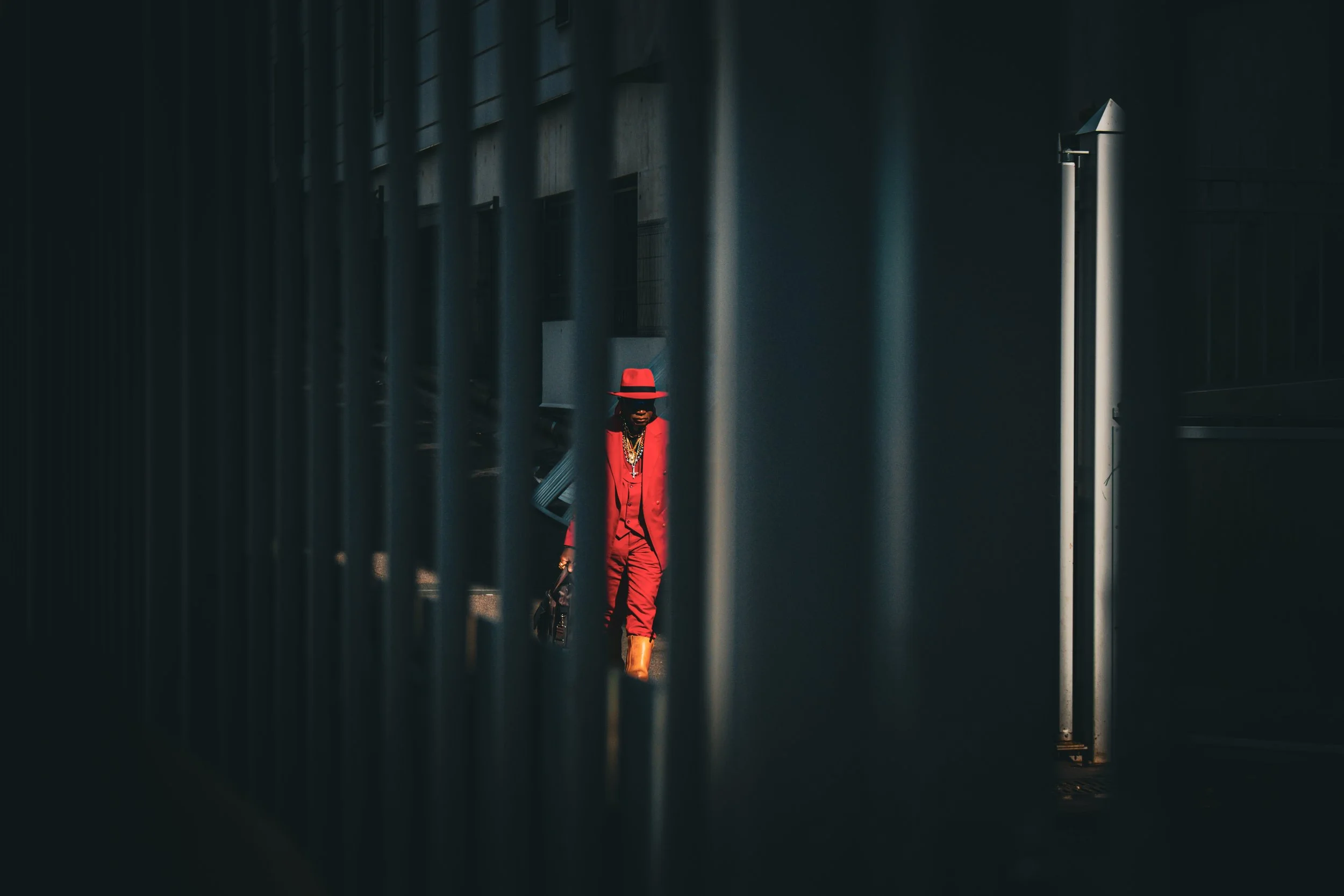 A person dressed in a red suit, red hat, and brown boots, walking behind a dark metal fence, holding a small bag, with a wall and vertical light fixtures in the background.