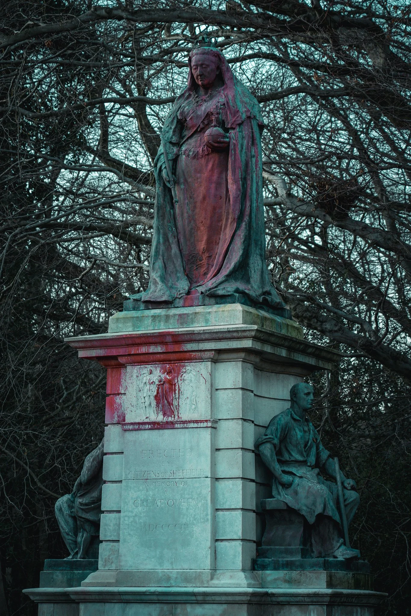 A bronze statue of Queen Victoria on a stone pedestal, with two seated figures on either side, surrounded by leafless trees.