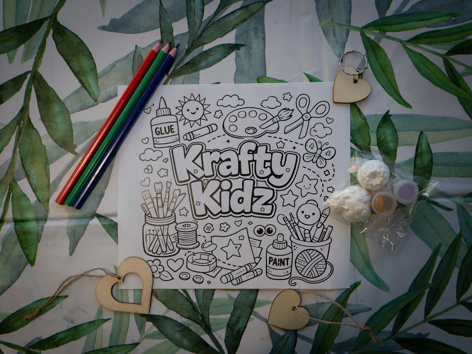 Black and white coloring page with the text 'Krafty Kidz' surrounded by art supplies like paintbrushes, glue, scissors, buttons, and decorative items on a leafy background with wooden heart-shaped accents.