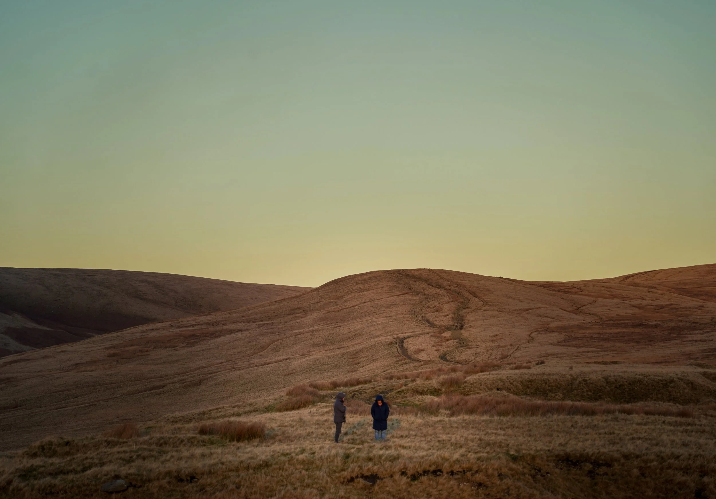 Two people standing in a vast, hilly landscape with dry grass, under a gradient sky during sunset or sunrise.