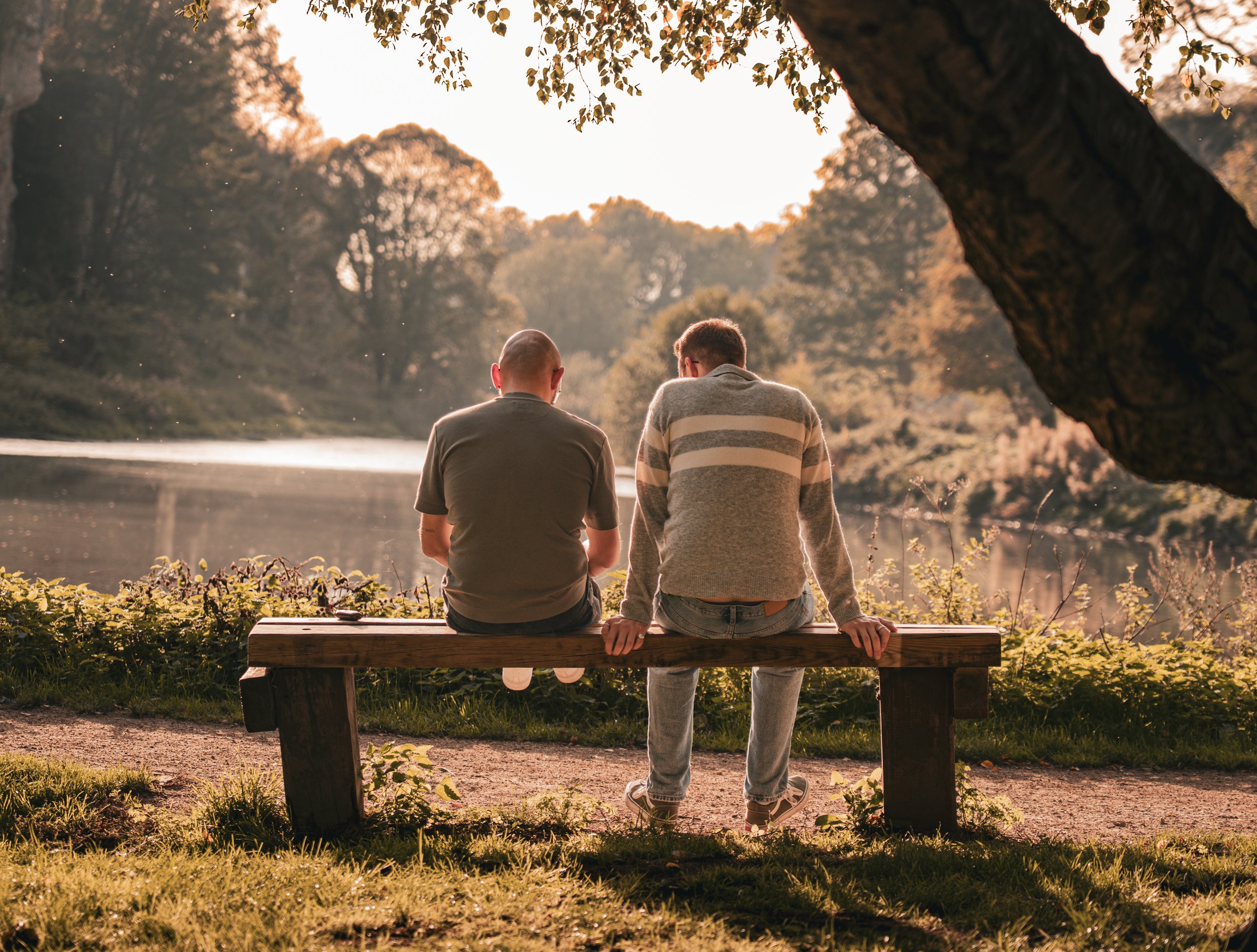 Two men sit on a wooden bench by a lakeside during sunset, with trees and a large rock in the background.