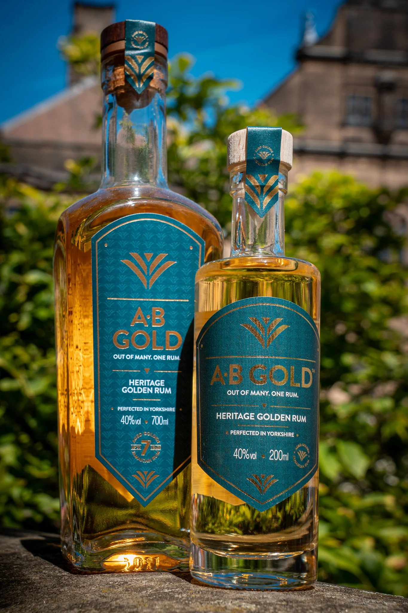 Two bottles of AB Gold Heritage Golden Rum outdoors on a surface, with a blue sky and trees in the background. The bottles are transparent with blue and gold labels, showcasing the golden rum inside.