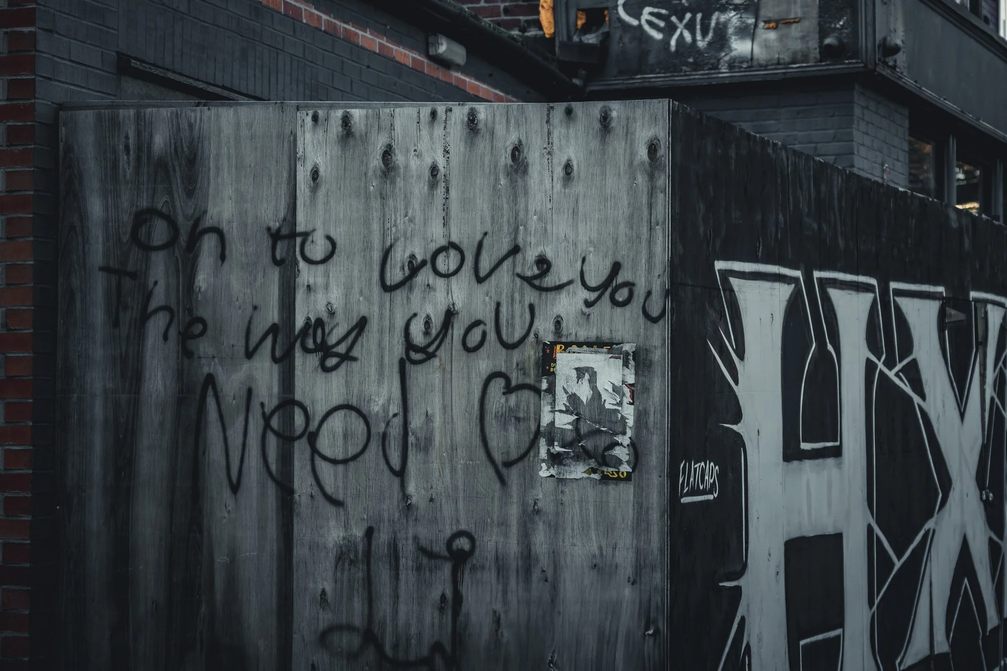 Graffiti-covered wooden wall with a handwritten message in black spray paint that says, "on to love you, the way you need". There is also a smaller tag that says "FLATCAPS" and a small poster or sticker partially torn.