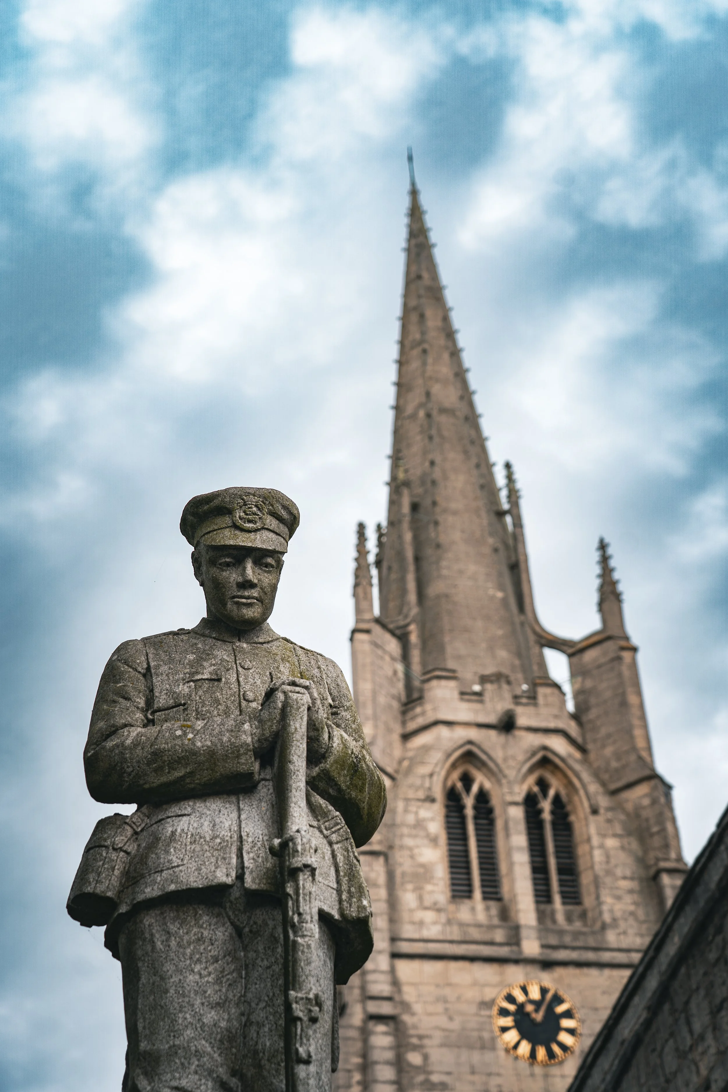 Stone statue of a soldier with hands clasped, in front of a historic church with a tall spire and clock tower, under cloudy sky.