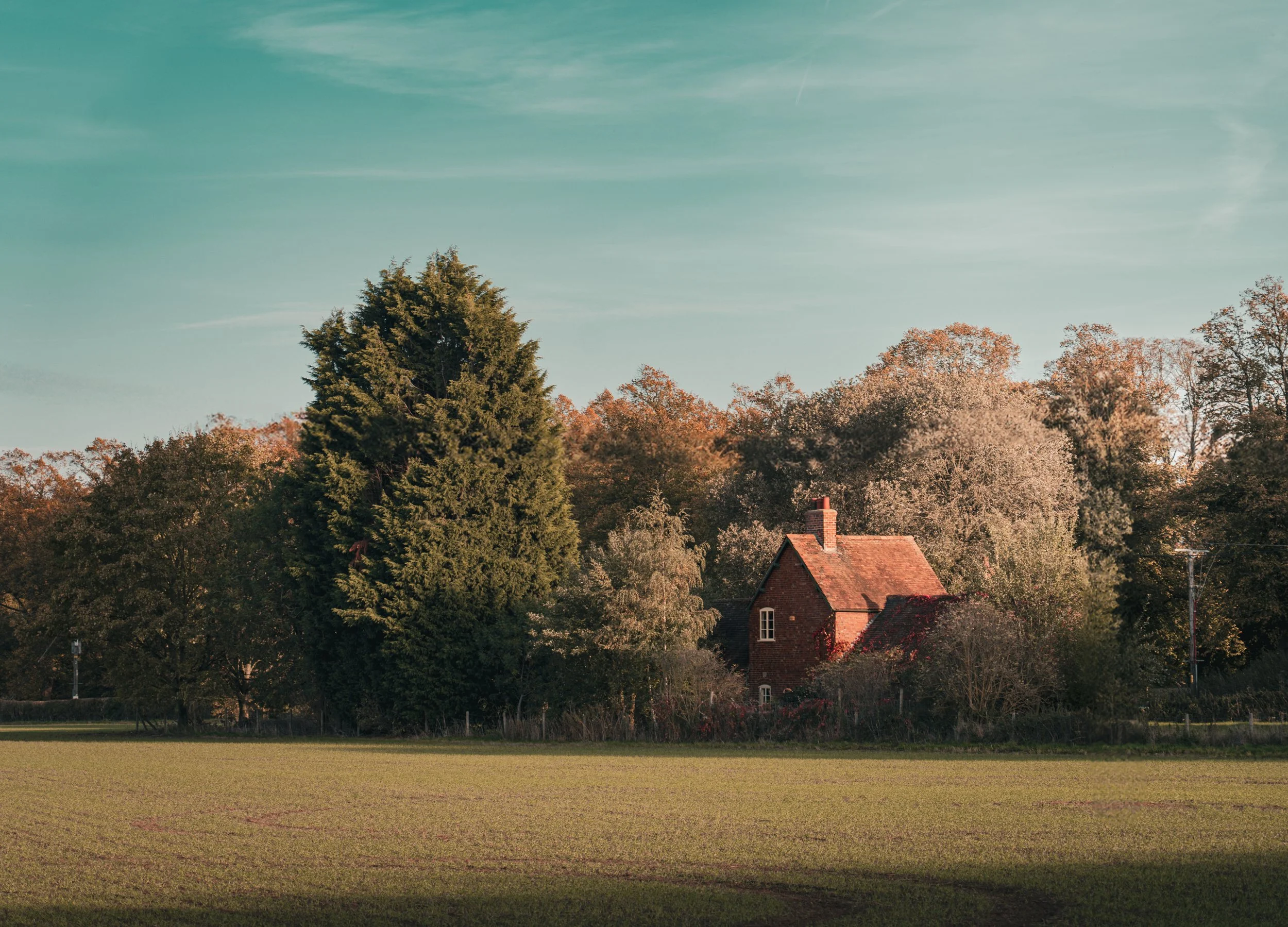 A red brick house surrounded by trees with fall foliage, situated behind an open grassy field under a blue sky.